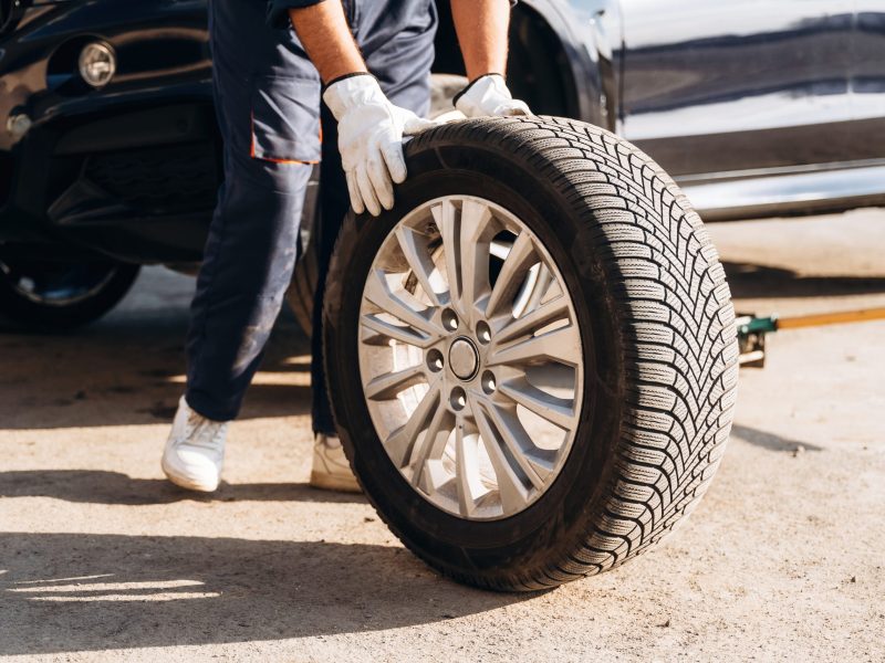 Close up view, with new tire. Mechanic is with the car outdoors.